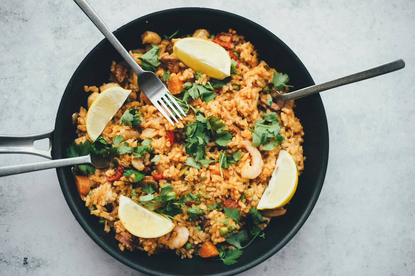 Overhead view of a shared pan of paella-style rice with lemon wedges, herbs and two forks